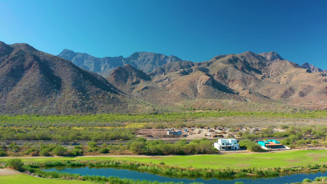Aerial view in Loreto, Baja California Sur where blue water crosses the foreground, green vegetation fills the middle, and small buildings sit below dry mountains with no visible people.