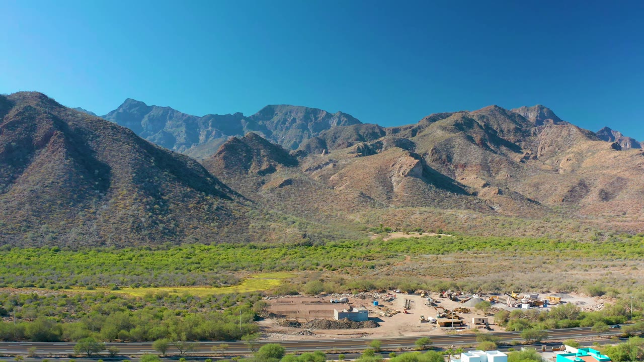 Aerial view in Loreto, Baja California Sur, with the small settlement and dirt road near the lower edge, green brush through the valley, and layered brown mountains beyond, with no visible people.
