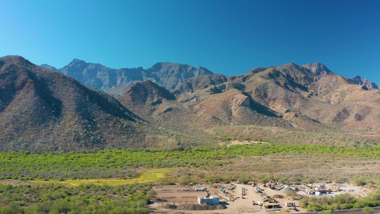 A slightly lower-looking aerial frame over Loreto, Baja California Sur, with green vegetation spanning the midground, a dirt settlement area near the bottom, and large desert mountains filling the background, with no visible people.