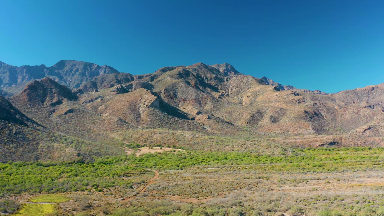 A wide Loreto valley frame shows the dirt road bending through green scrub and pale dry ground, with layered brown mountains across the horizon and no visible people.