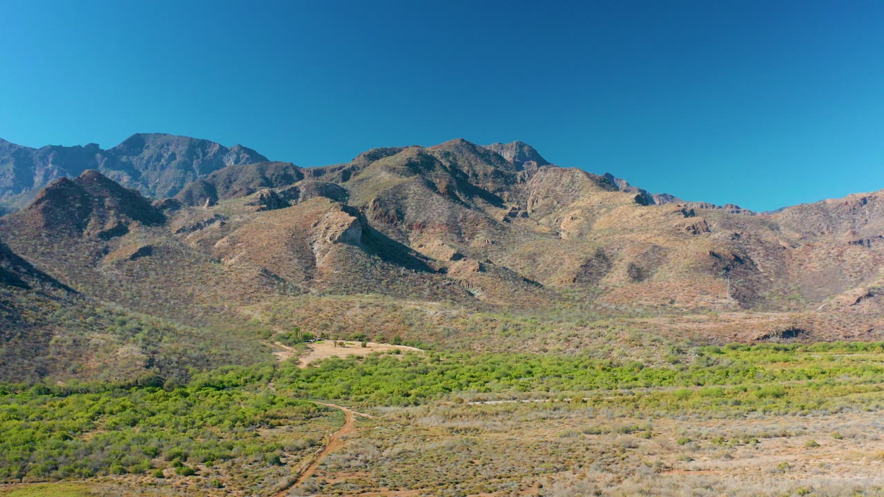 Opening view of rocky mountains and a green valley in Loreto, Baja California Sur, with scrubby foreground terrain and no visible people.