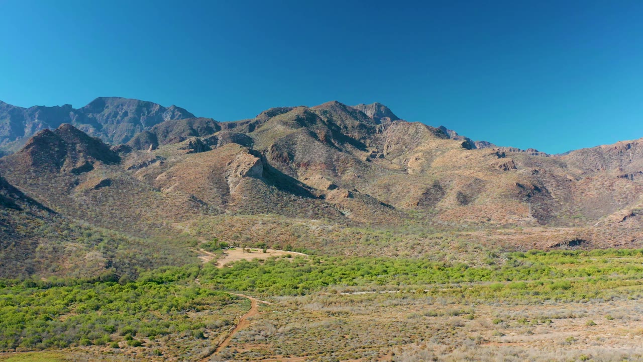 Wide aerial view in Loreto, Baja California Sur, showing tan rocky slopes, green valley vegetation, a pale dirt track, and no visible people.