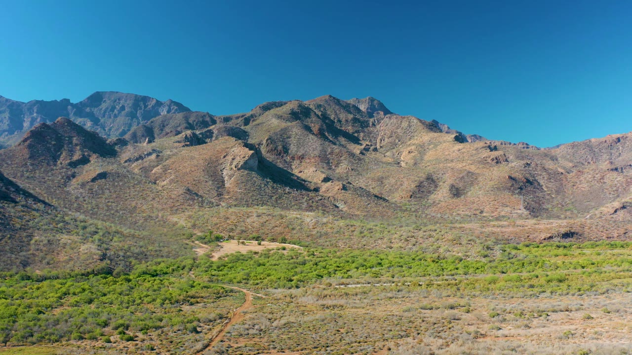 High view over Loreto, Baja California Sur, with a dark mountain shoulder on the left, lighter rocky hills to the right, green valley growth, and no visible people.