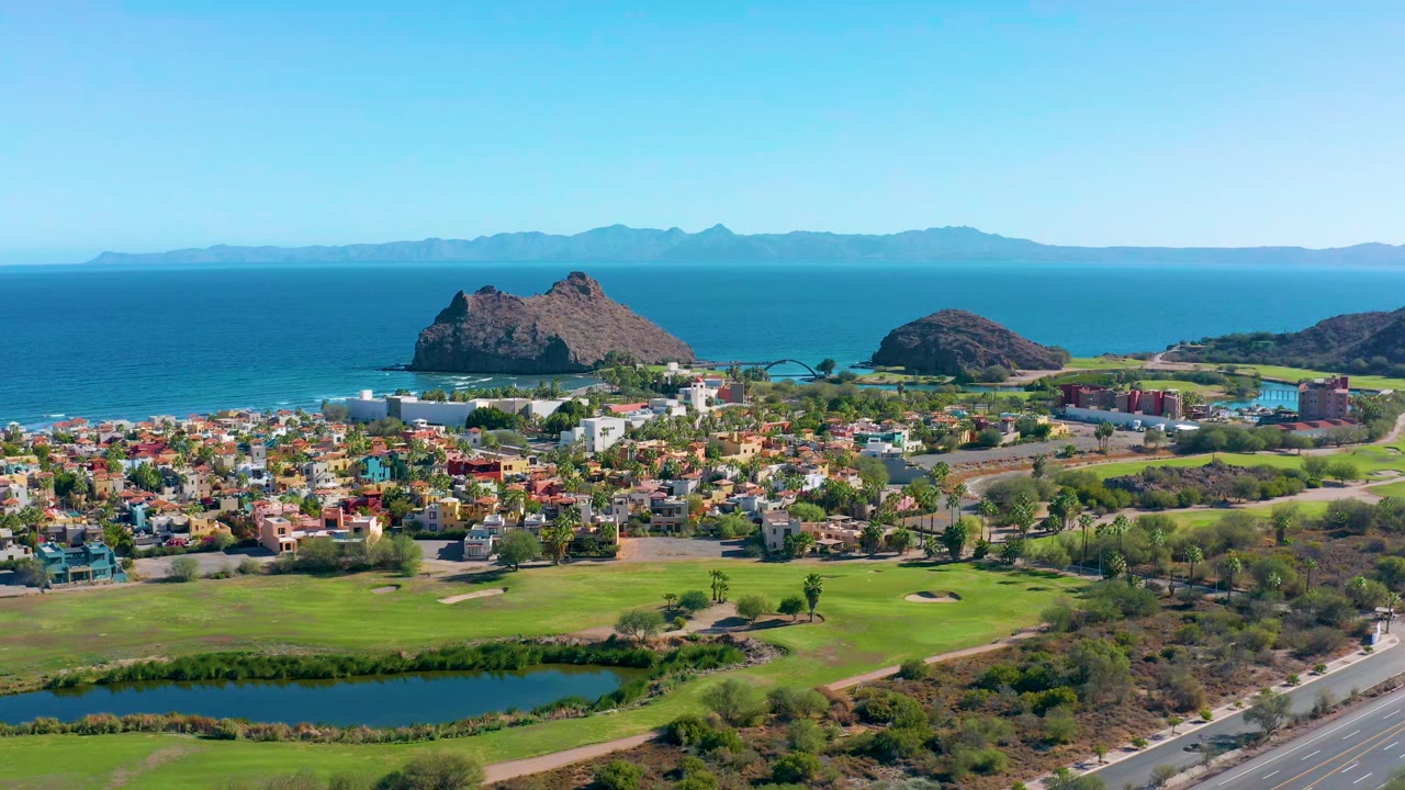 Opening aerial frame over Loreto, Baja California Sur with a golf course pond in the foreground, colorful homes in the midground, blue ocean and rocky hills beyond, and no visible people.
