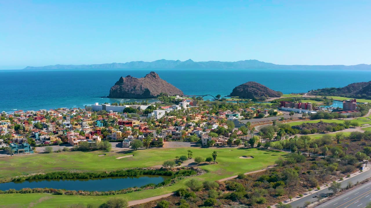 Wide aerial of homes beside a green golf course with ocean and rocky hills behind.