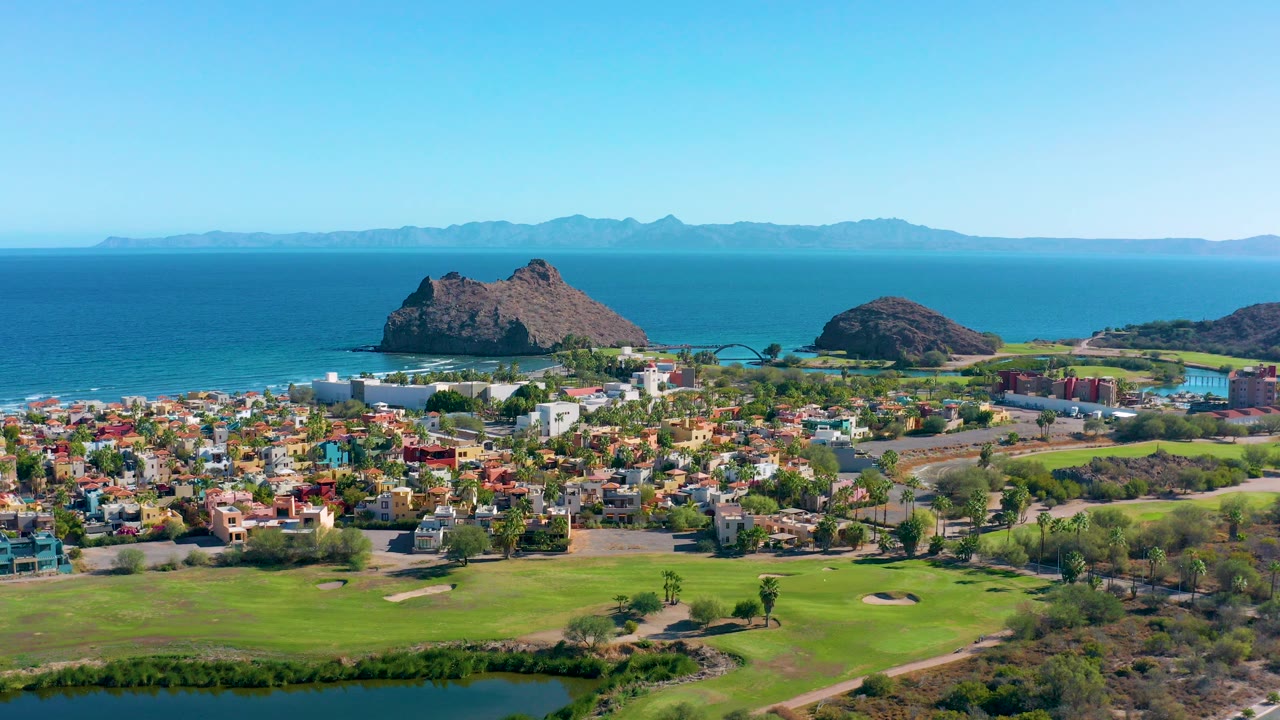 Wide aerial view over Loreto, Baja California Sur, with green fairways, residential blocks, blue ocean, rocky offshore outcrops, distant land on the horizon, and no visible people.