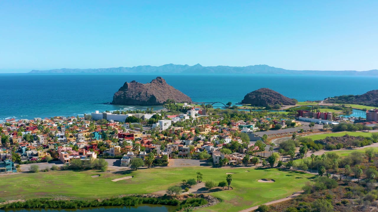 High aerial view of Loreto with a broad golf course foreground, dense residential area, a pair of rocky coastal outcrops, blue sea, distant landforms, and no visible people.