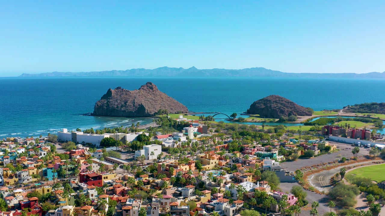 Opening aerial frame over Loreto, Baja California Sur, with colorful low-rise buildings below, rocky islands offshore, blue ocean, distant mountains, and no visible people.