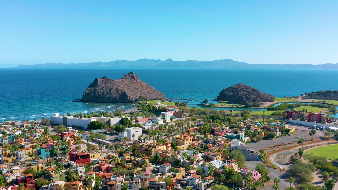 Aerial view over Loreto showing the town grid and palms in the lower frame, a white coastal building, rocky islands near shore, blue water, distant mountains, and no visible people.