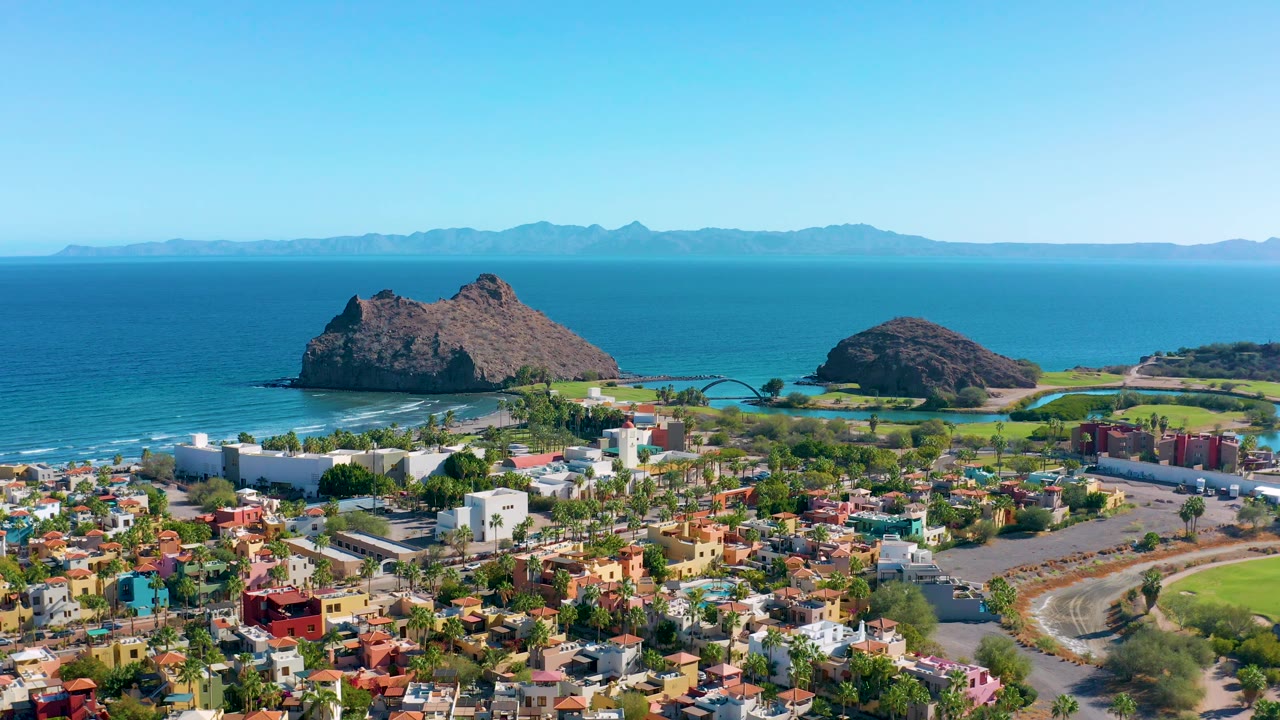 Loreto aerial frame with dense colorful buildings and a curved road in front, green coastal land behind them, two rocky offshore landforms, blue ocean, and no visible people.