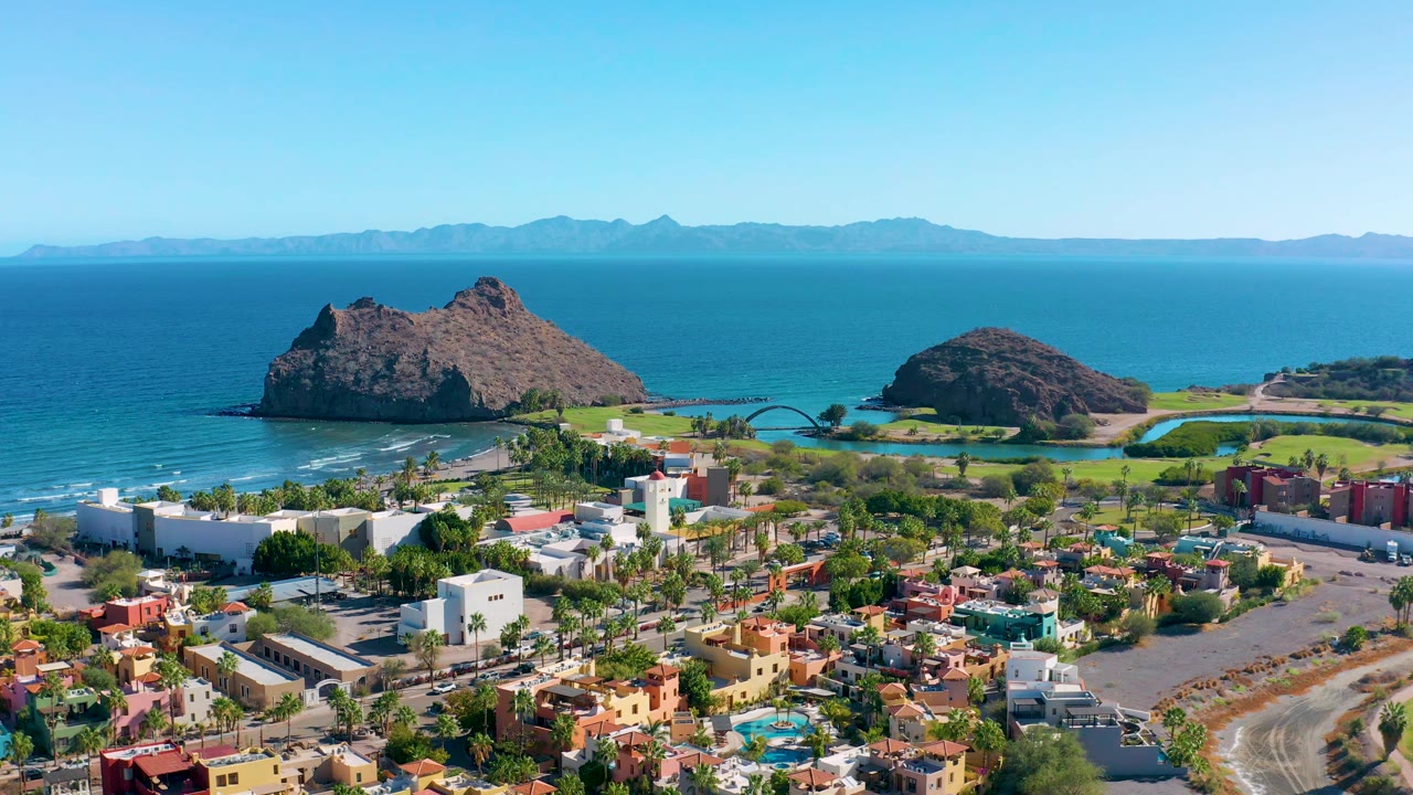 Wide aerial coastline with town blocks, palms, beach, and rocky islands offshore.