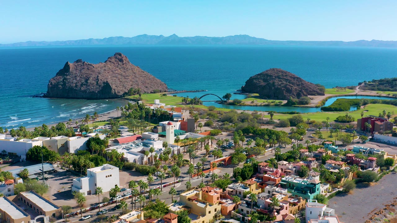 Wide aerial frame over Loreto, Baja California Sur, with colorful low buildings, palm-lined streets, blue ocean, two offshore rock formations, distant mountains, and no visible people.