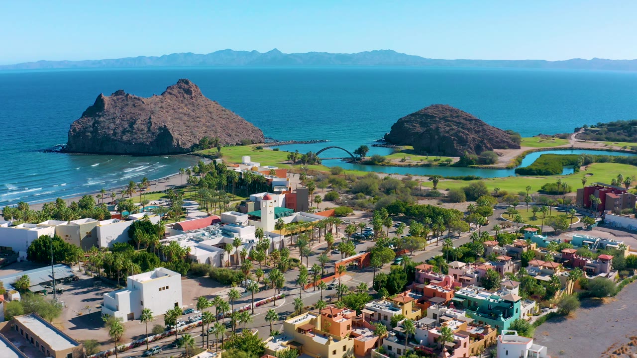 Loreto aerial frame with town blocks and palms in the lower view, green shoreline grounds at center, two brown rock formations in blue water, distant mountains, and no visible people.