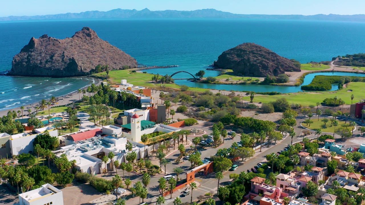 Aerial view of rocky coastline, blue ocean, bridge, buildings, roads, palms, and parked cars.
