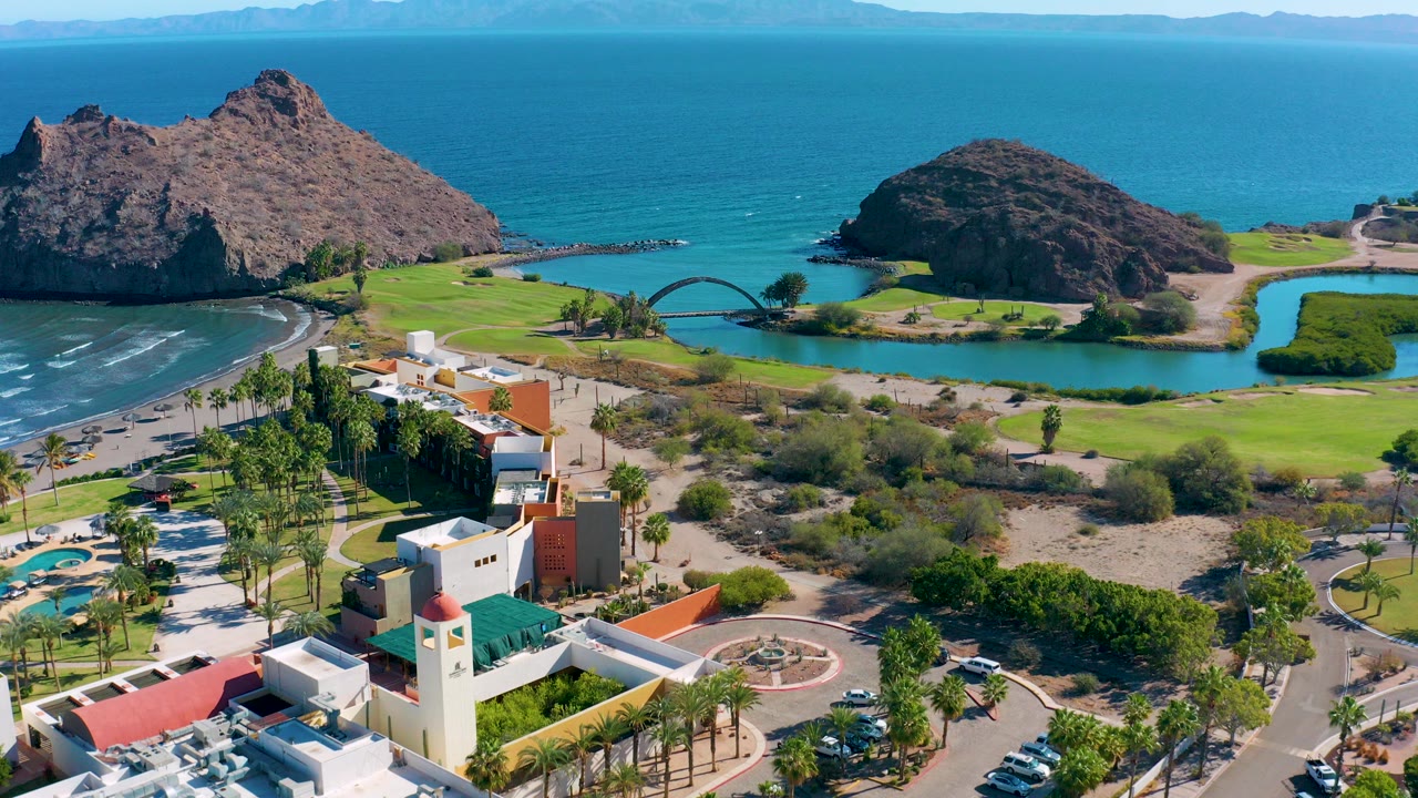 Aerial frame over Loreto, Baja California Sur showing resort buildings and palms in the foreground, a green golf course and lagoon with an arched bridge in the middle, rocky coastal formations, open ocean, and no visible people.