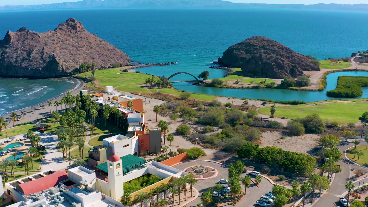 Drone view of resort complex, palms, lagoon bridge, golf greens, rock formations, and ocean.