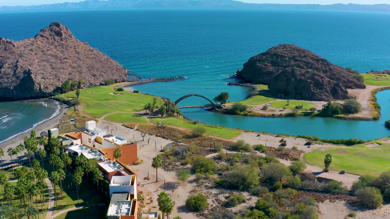 A second aerial moment in Loreto, Baja California Sur keeps the bridge centered between the lagoon and green grass, with rocky hills framing the blue ocean and no visible people.