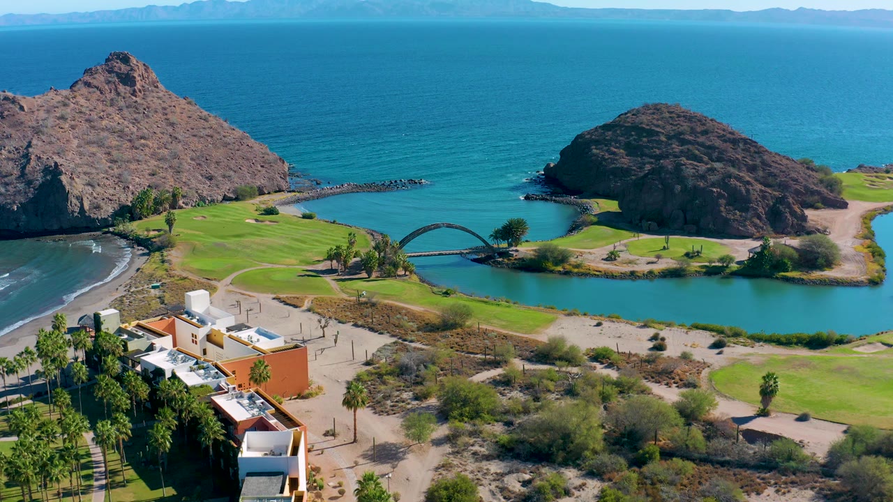 Aerial view in Loreto, Baja California Sur with the lagoon stretching across the lower frame, the arched bridge near center, rocky hills at left and right, and no visible people.