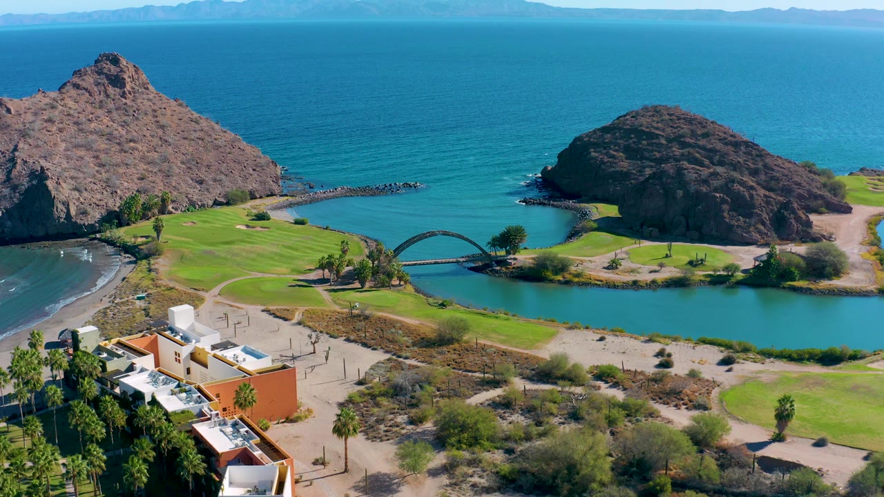 Mid-clip aerial frame over Loreto, Baja California Sur showing small white wave lines on the beach, turquoise lagoon water, the arched bridge, green grass, rocky hills, and no visible people.