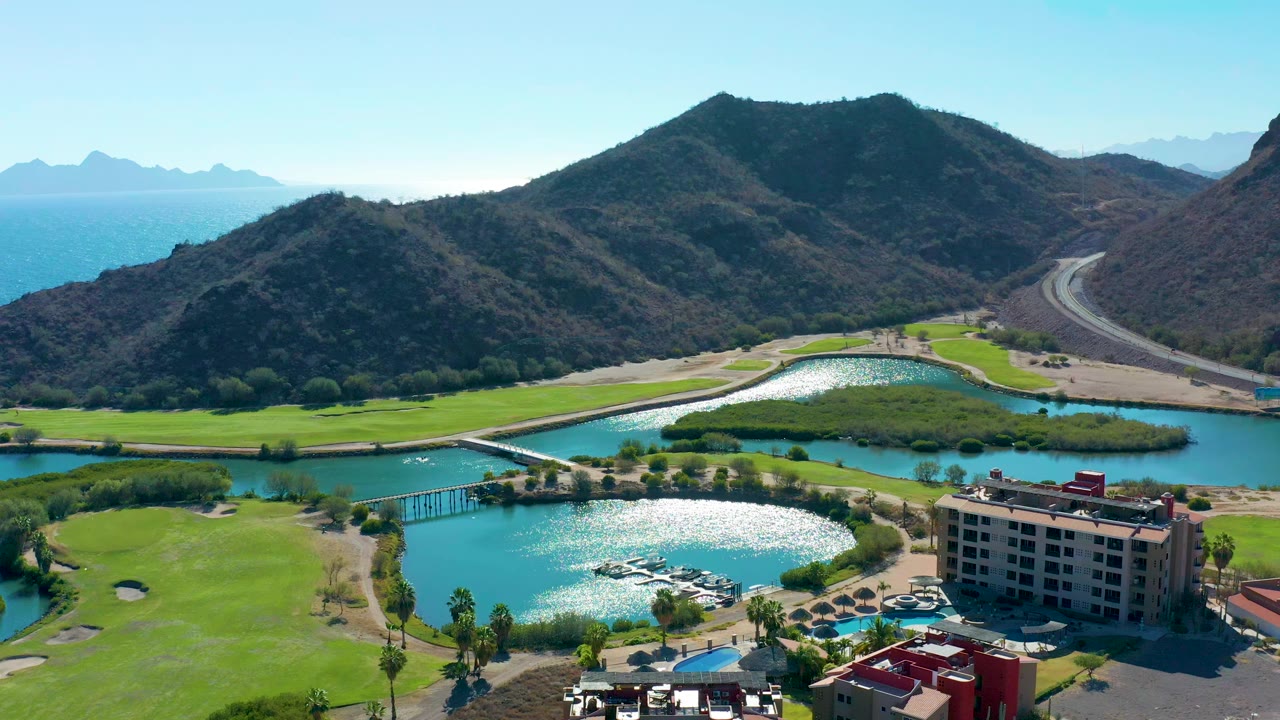 Aerial frame in Loreto, Baja California Sur showing turquoise lake channels, bright golf greens, a resort building, a road at the mountain base, distant coastline, and no visible people.