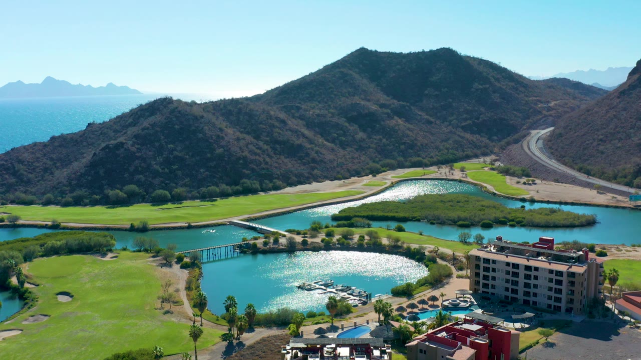 Drone view of blue lake water, golf greens, small bridges, resort structures, road, and hills.