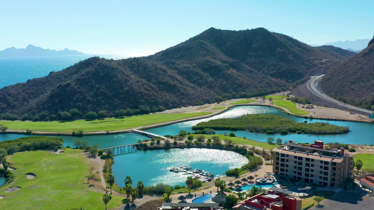 Loreto aerial view with the hotel slightly lower in frame, sun flashing on lagoon water, green fairways around the shoreline, a bridge at left, dry mountains, road, ocean, and no visible people.