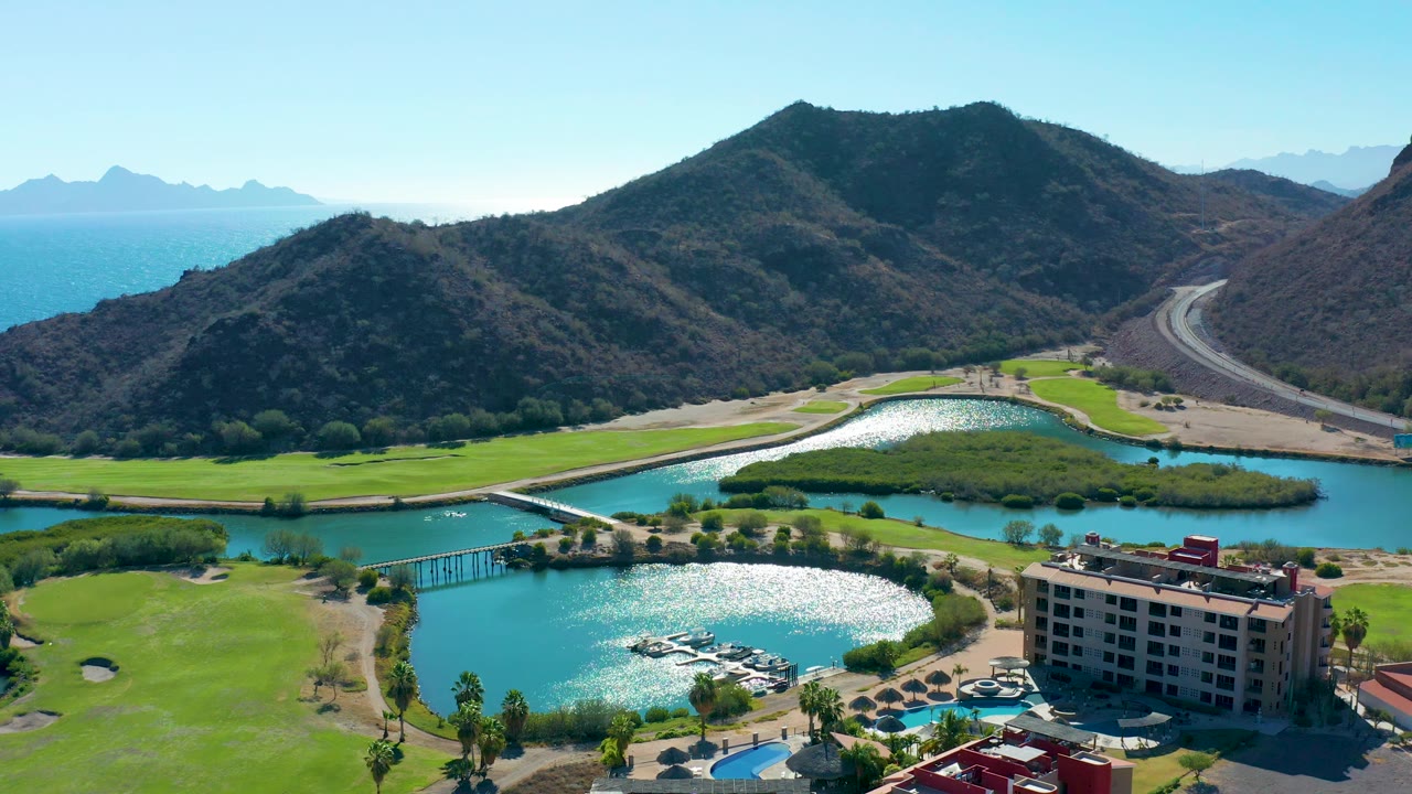 Loreto aerial frame with the lagoon sparkle shifted across the water, the golf course wrapping the shore, the hotel at lower right, a bridge and boats below, mountains behind, ocean at left, and no visible people.