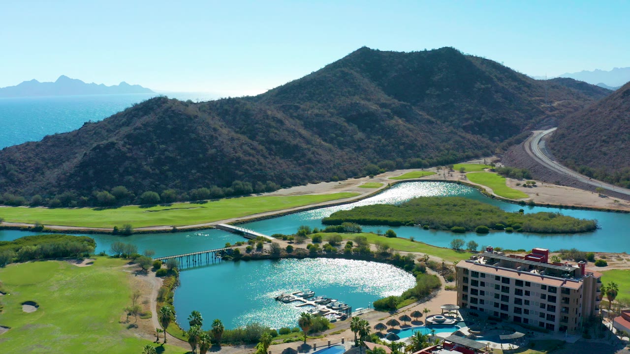 Aerial view of a golf course, lake, building, road, mountains, and blue coastal water.
