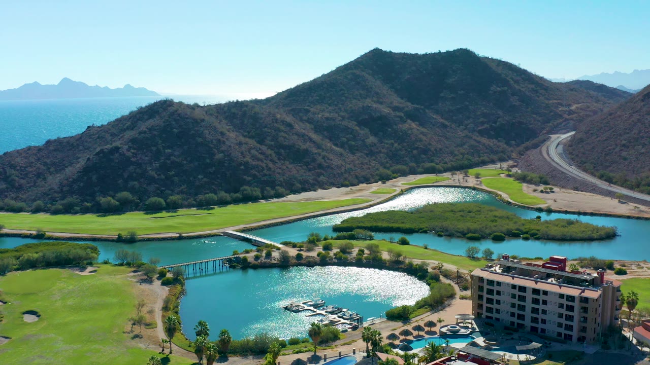 Wide aerial frame over Loreto with green fairways and turquoise water in the foreground, a small dock area near the lower center, mountains behind, a hillside road at right, and no visible people.