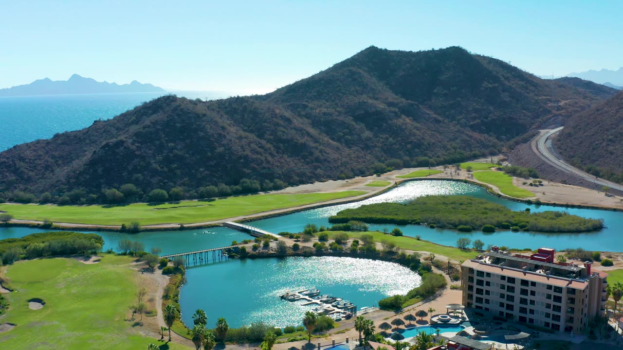 Aerial landscape in Loreto showing reflective lake water, golf greens, a bridge across the water, the edge of a multi-story building, a winding road on the hillside, mountains, and no visible people.