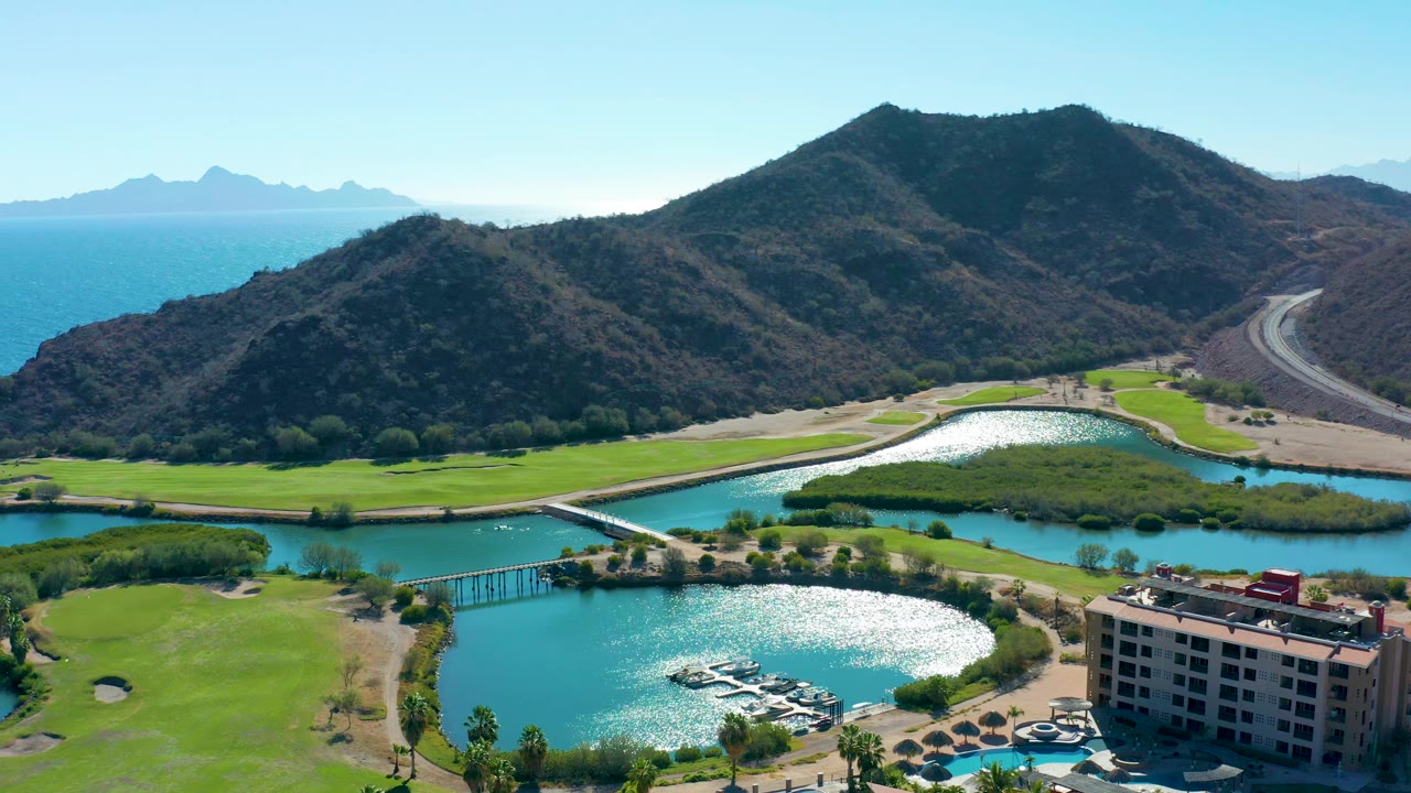 Aerial frame over Loreto with golf course grass and water channels across the lower half, dark mountain slopes filling the center, blue sea visible beyond the left ridge, and no visible people.