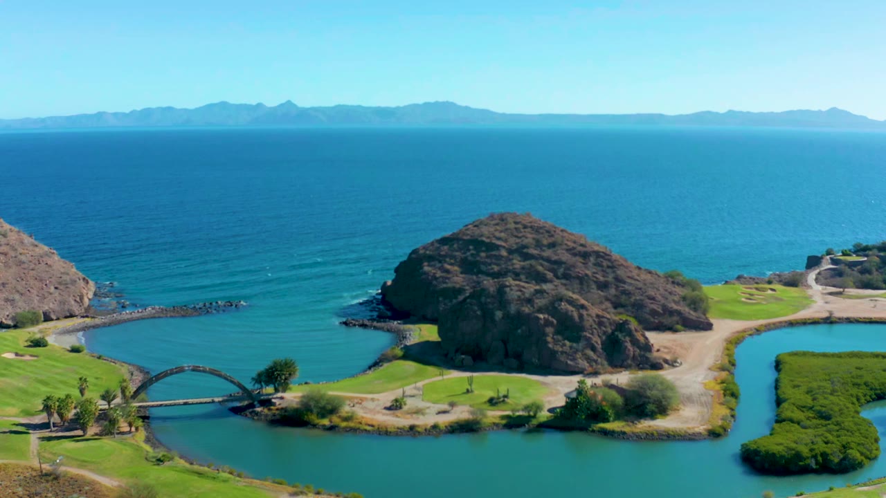 Loreto, Baja California Sur shoreline frame with the rocky hill centered, the bridge crossing water in the lower left, ocean waves in the background, and no visible people.