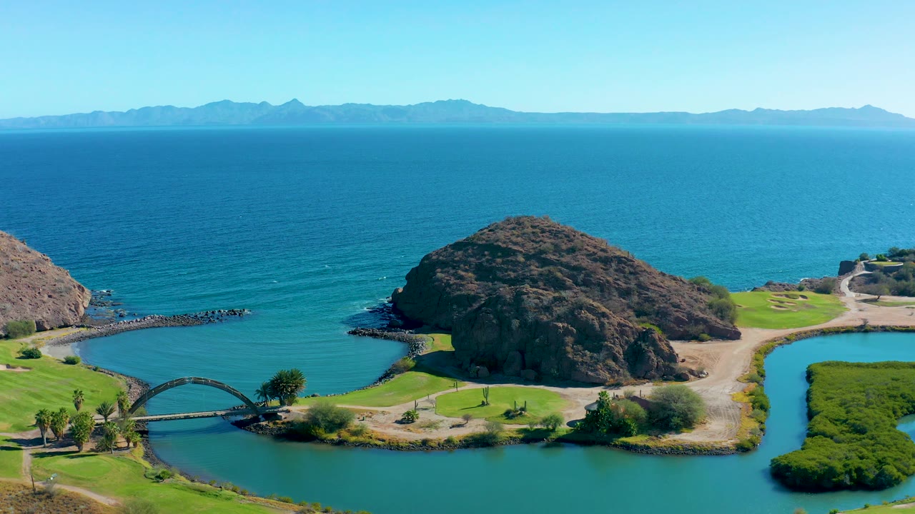 Loreto, Baja California Sur coastal landscape with green grounds split by a narrow channel, a small bridge, a rocky hill, blue ocean, distant mountain ridges, and no visible people.