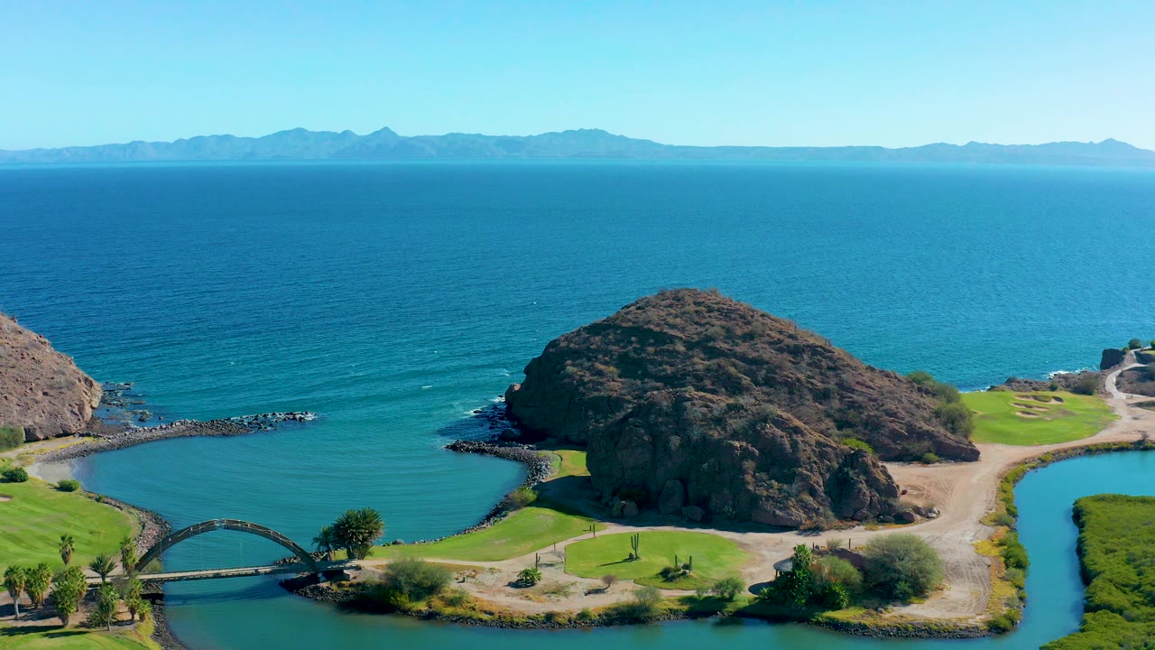 Aerial view in Loreto, Baja California Sur showing a rocky cove, small bridge, green lawn edges, blue ocean, distant mountains, and no visible people at the opening frame.