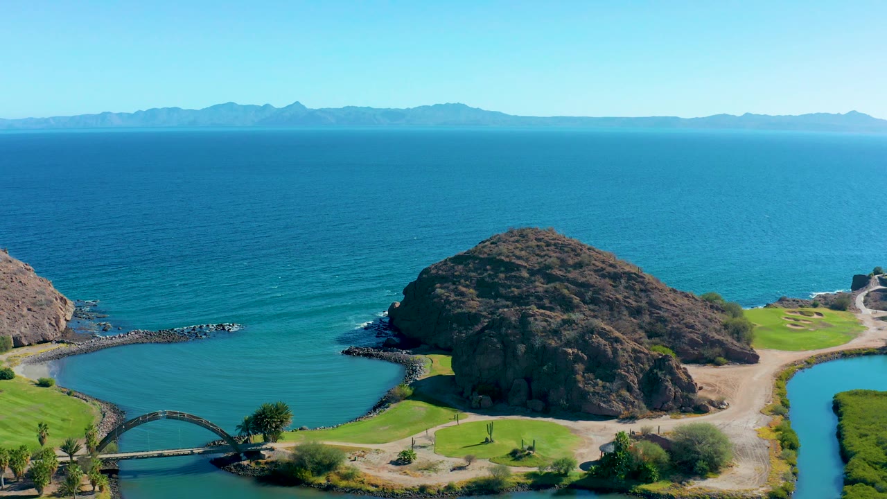 Aerial view in Loreto, Baja California Sur showing the cove from a similar high angle, with the rock formation near center, ocean ripples beyond, green patches, and no visible people.