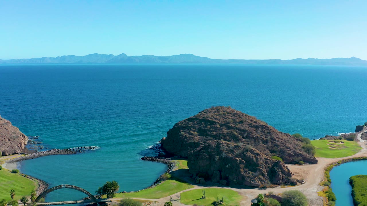 High view over a blue coastal inlet with rocky mound, green areas, and distant mountains.