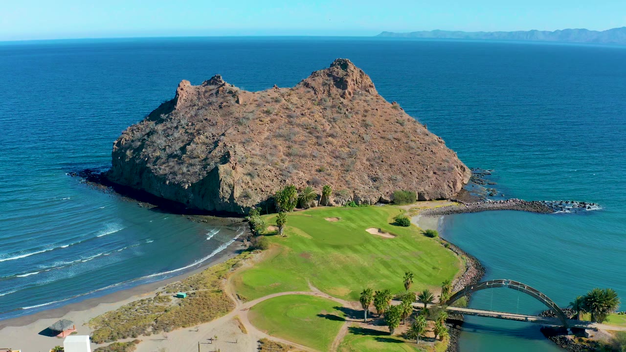 Aerial frame in Loreto, Baja California Sur showing the rock headland slightly centered over the green field, with the curved bridge at the lower right and no visible people.