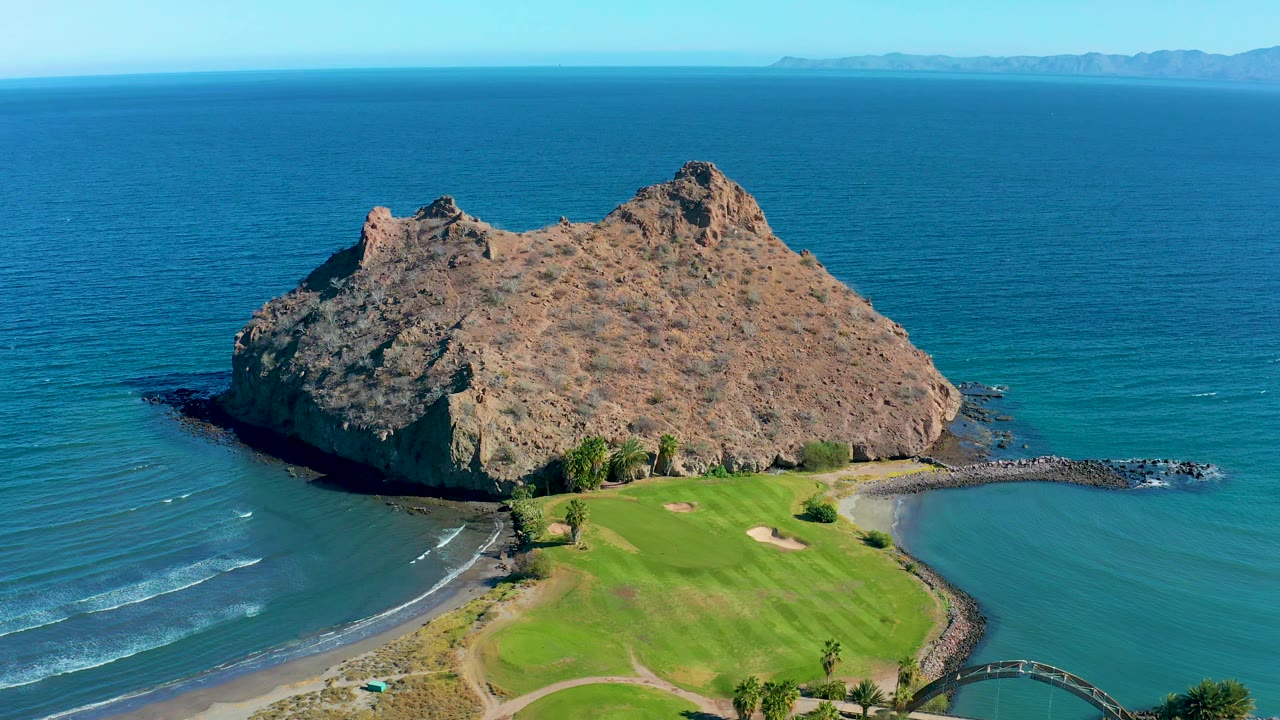 Aerial frame from Loreto, Baja California Sur, Mexico, with the rocky island centered above the golf green, small white wave lines on the left water, and no visible people.