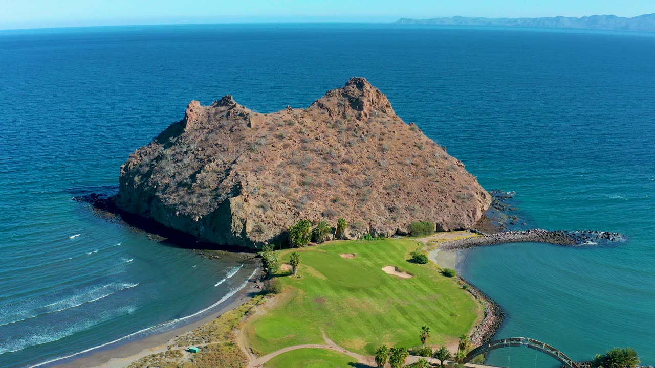 Aerial view in Loreto, Baja California Sur, Mexico, with the rocky island slightly right of center, the golf green below it, blue ocean across the background, and no visible people.