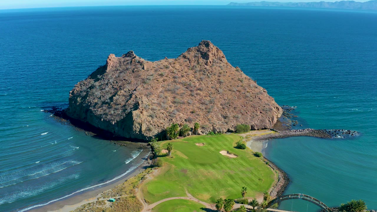 Aerial frame from Loreto, Baja California Sur, Mexico, showing the rocky island above the golf green, a curving beach at left, darker rocks along the right edge, and no visible people.