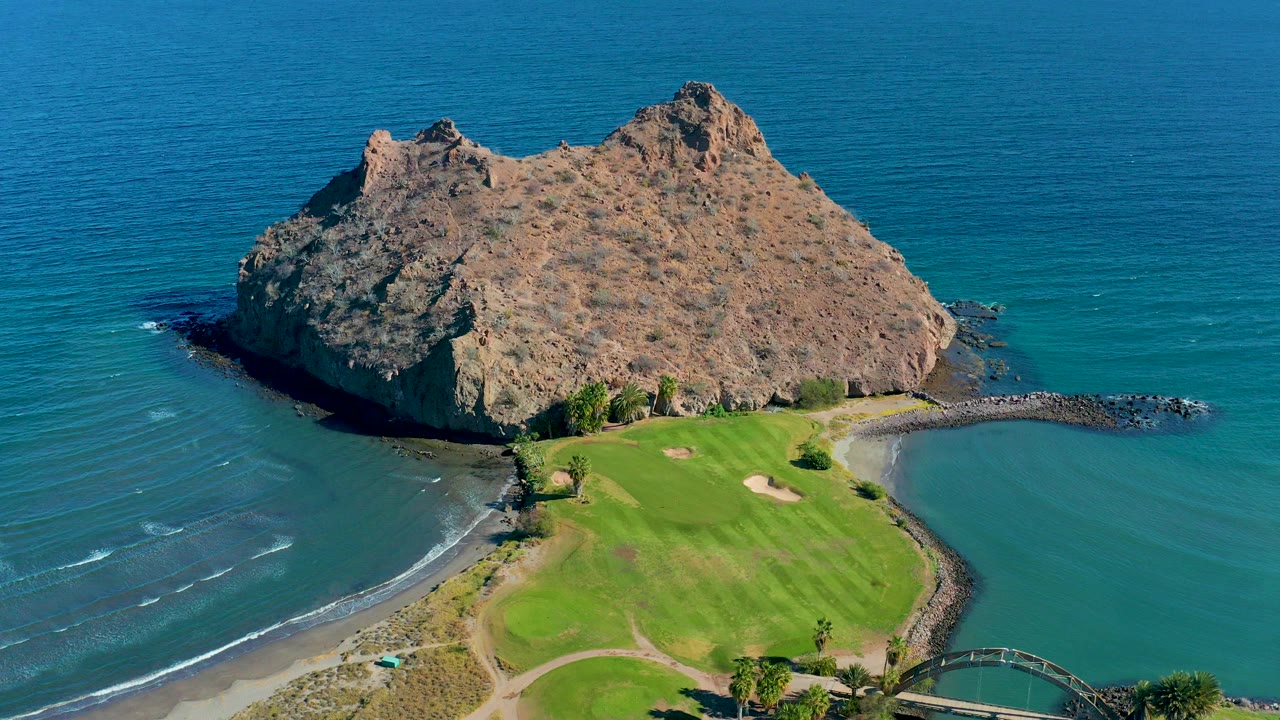 Aerial view in Loreto, Baja California Sur showing a rocky island above turquoise water, a green golf course, beach edge, and no visible people.