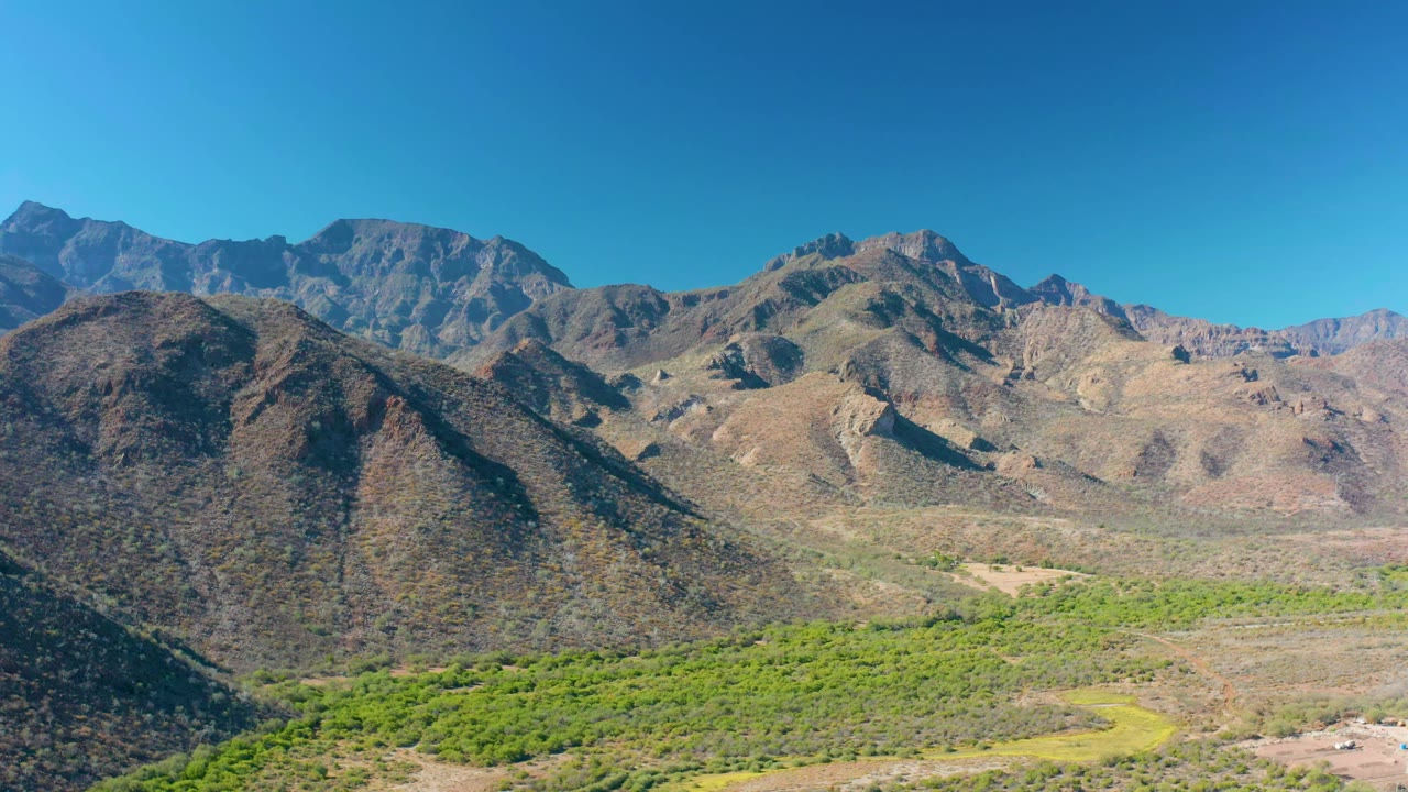 Early frame of the Loreto mountain valley, with a dark rounded hillside on the left, tan ridges across the center, green vegetation below, and no visible people.