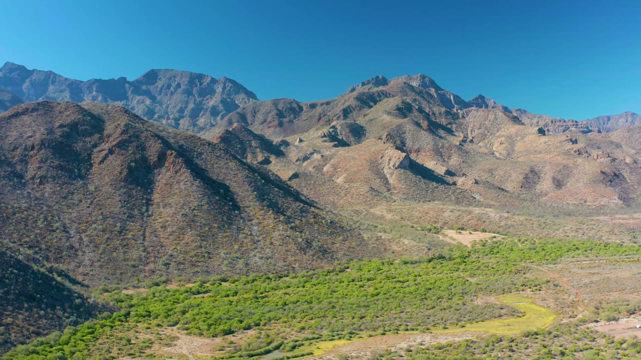 Aerial frame in Loreto, Baja California Sur showing brown ridges set behind a dense green strip of valley vegetation, with no visible people.