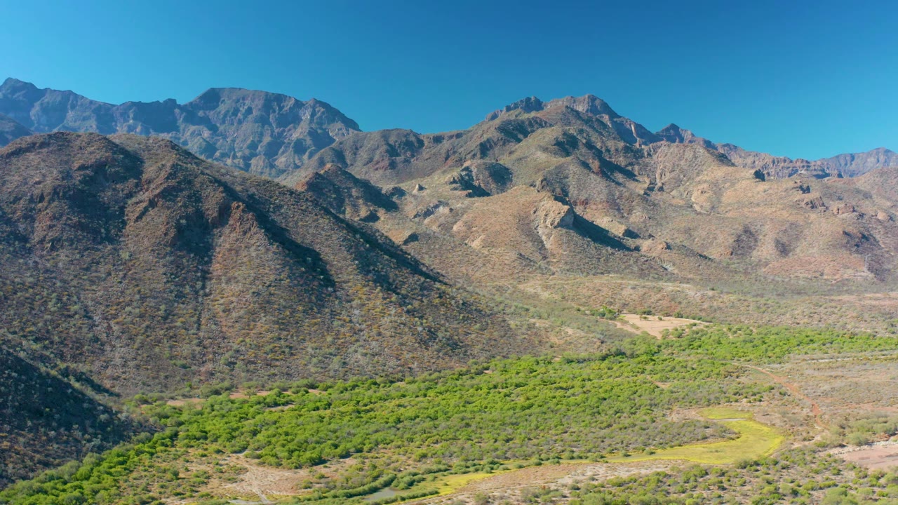 High landscape view of Loreto mountains with a bright green valley floor, dry rocky terrain, and a cloudless blue sky, with no visible people.
