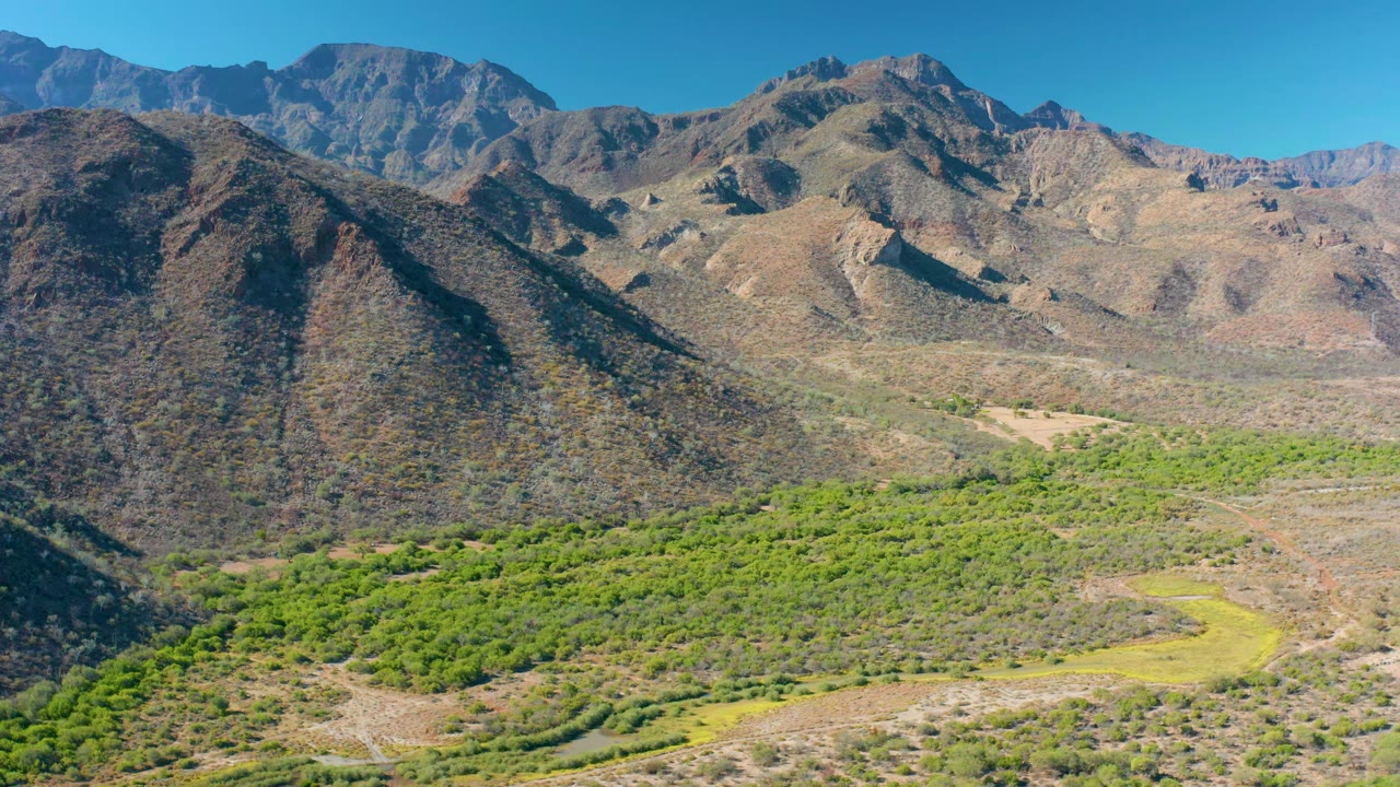 A wide aerial view in Loreto, Baja California Sur shows the green valley band more centrally below rugged brown hills and distant ridges, with no visible people.