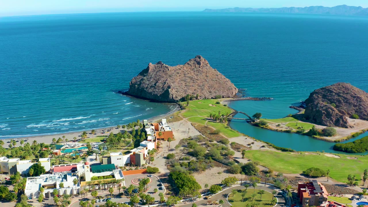 Aerial view over Loreto, Baja California Sur showing blue ocean, a lagoon, green shoreline grounds, tan rocky outcrops, coastal buildings, distant mountains, and no visible people.