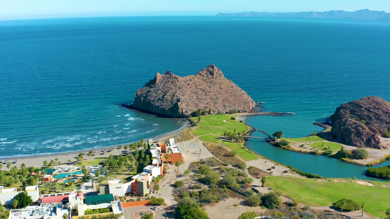 Loreto coastal aerial with small surf lines reaching the beach, a green lagoon beside manicured grass, resort buildings in the lower frame, and no visible people.