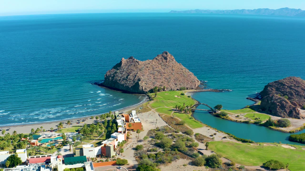 Aerial view over Loreto with the rocky outcrop slightly right of center, bright green shoreline grounds, lagoon water, beach vehicles at a distance, and no visible people.