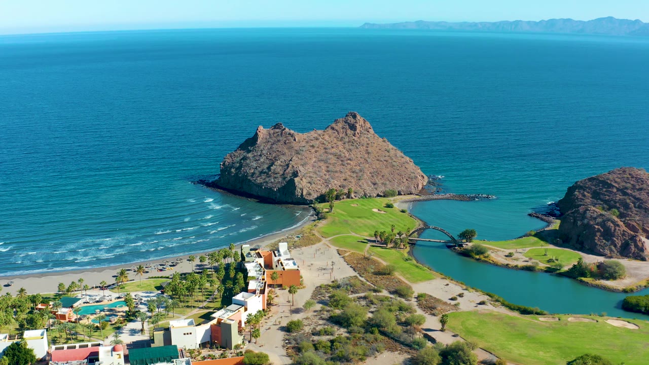 Loreto aerial frame with more open ocean above the rocky outcrop, pale surf bands on the beach, a green lagoon to the right, resort buildings below, and no visible people.