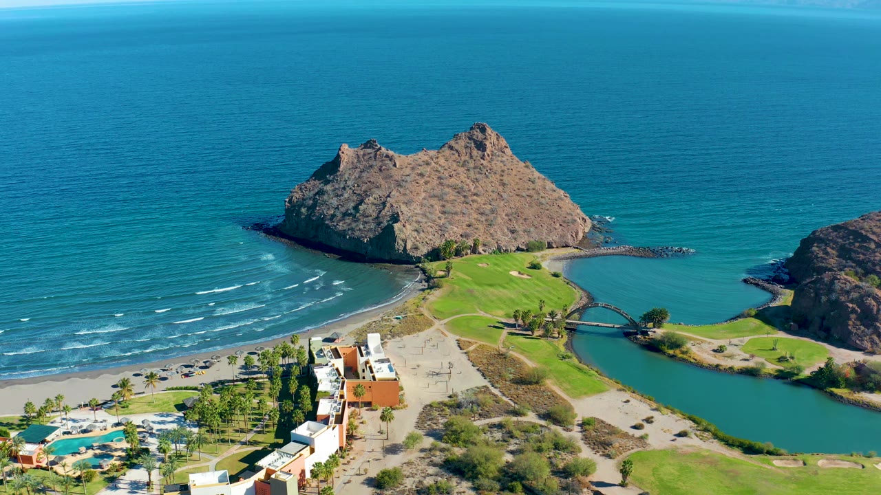 Drone view over ocean waves, rocky outcrop, beach, lagoon, bridge, and resort area.